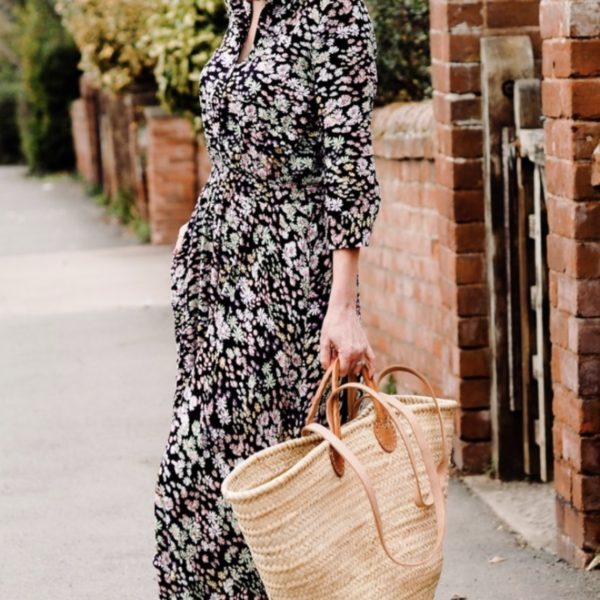 a lady wearing a spring dress walking down a quaint lane with a large french basket for her shopping